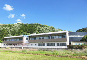 介護施設の外観。緑に囲まれた敷地に立つ2階建ての白とピンク色の建物。